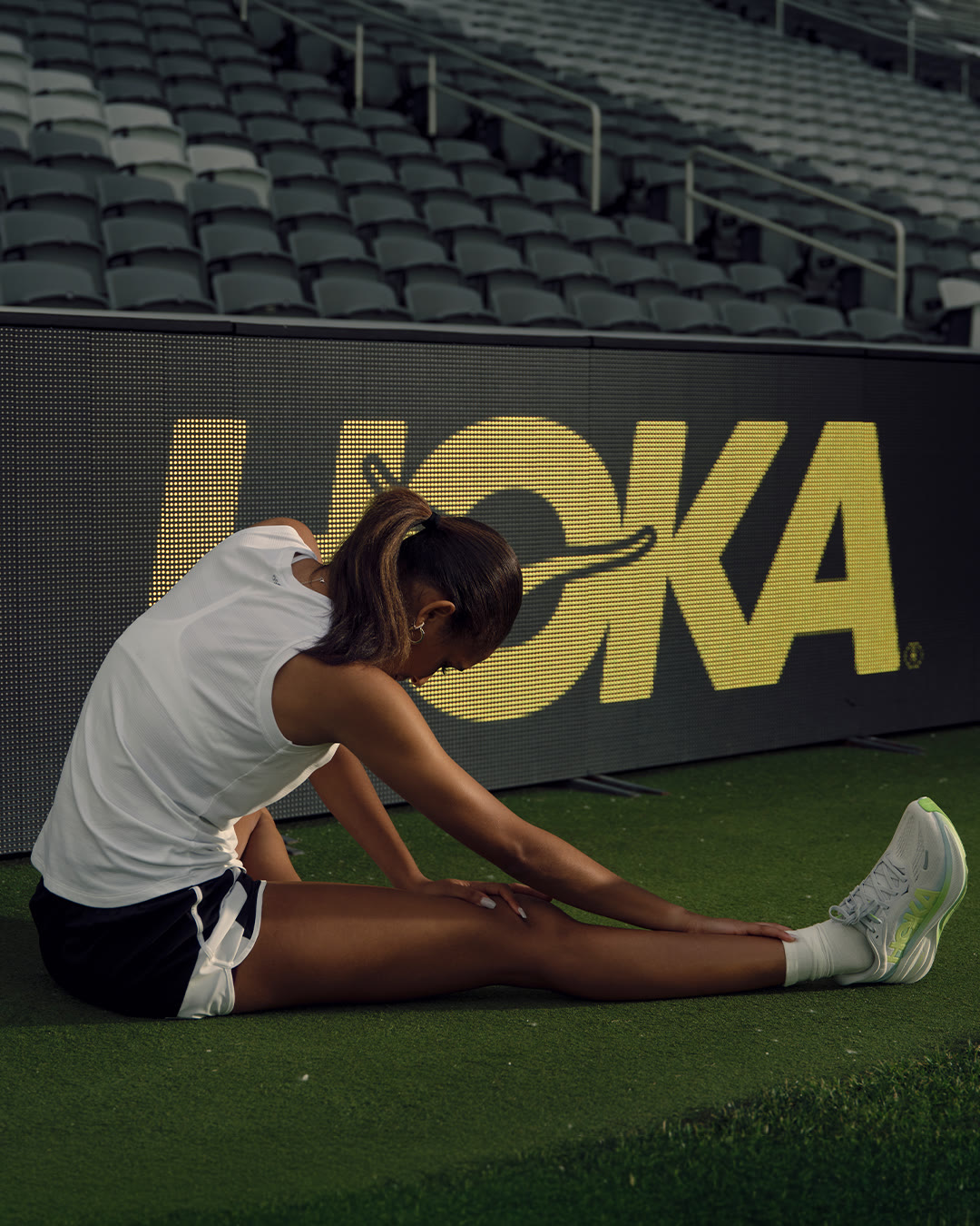 Anaiyah Robinson stretches on the turf in front of a glowing HOKA sign at dusk.
