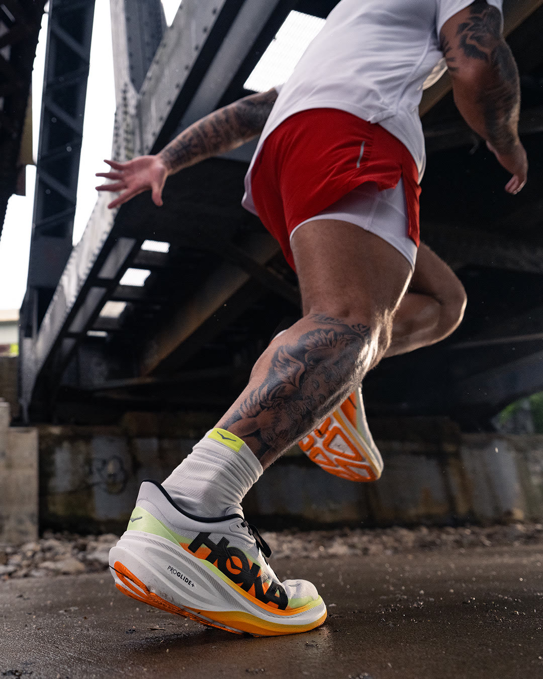 Close-up of a HOKA Clifton Pro mid-stride under a steel bridge.